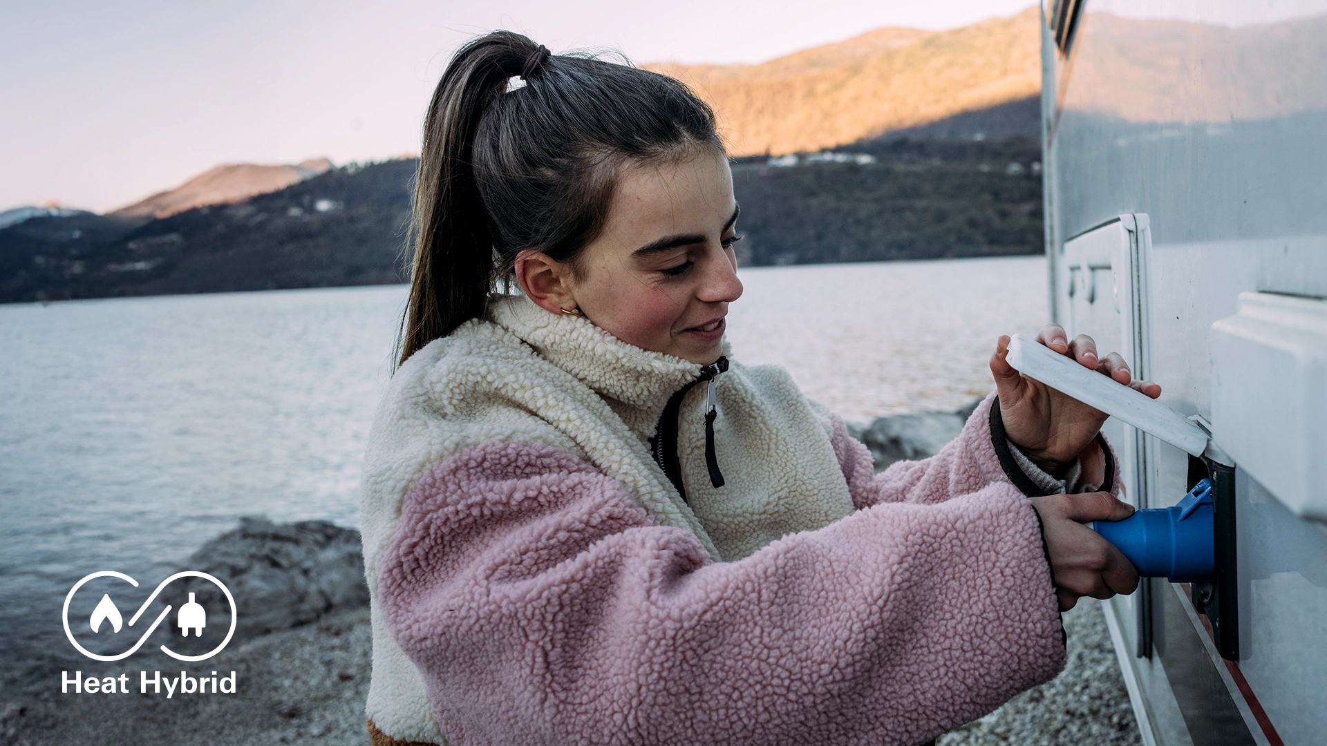 „Person steckt einen blauen Stromstecker in die Außensteckdose eines Wohnmobils, im Hintergrund ein See und Berge bei Tageslicht. Truma Logo ‚Heat Hybrid‘ unten links.“